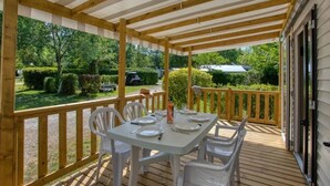 A covered outdoor dining area with a table set for four, surrounded by chairs, and a wooden deck. - Camping 3 étoiles - Piscine  - ccbfgee (Groléjac)