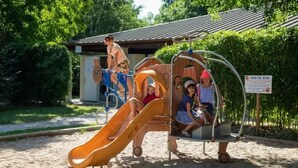 A playground with a slide, swings, and a small building in the background. - Camping 3 étoiles - Piscine  - ccbfgee (Groléjac)
