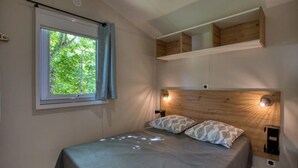 A bedroom with a bed, a window with curtains, a headboard with built-in lighting, and a wooden shelf above the bed. - Camping 3 étoiles - Piscine  - ccbfgee (Groléjac)