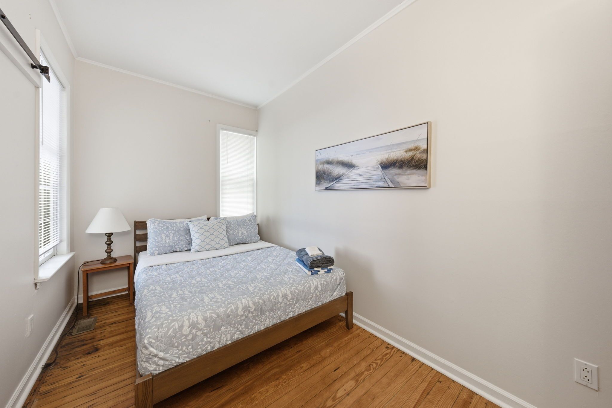 Un dormitorio con cama, mesita de noche, lámpara, ventana con persianas y un cuadro en la pared.