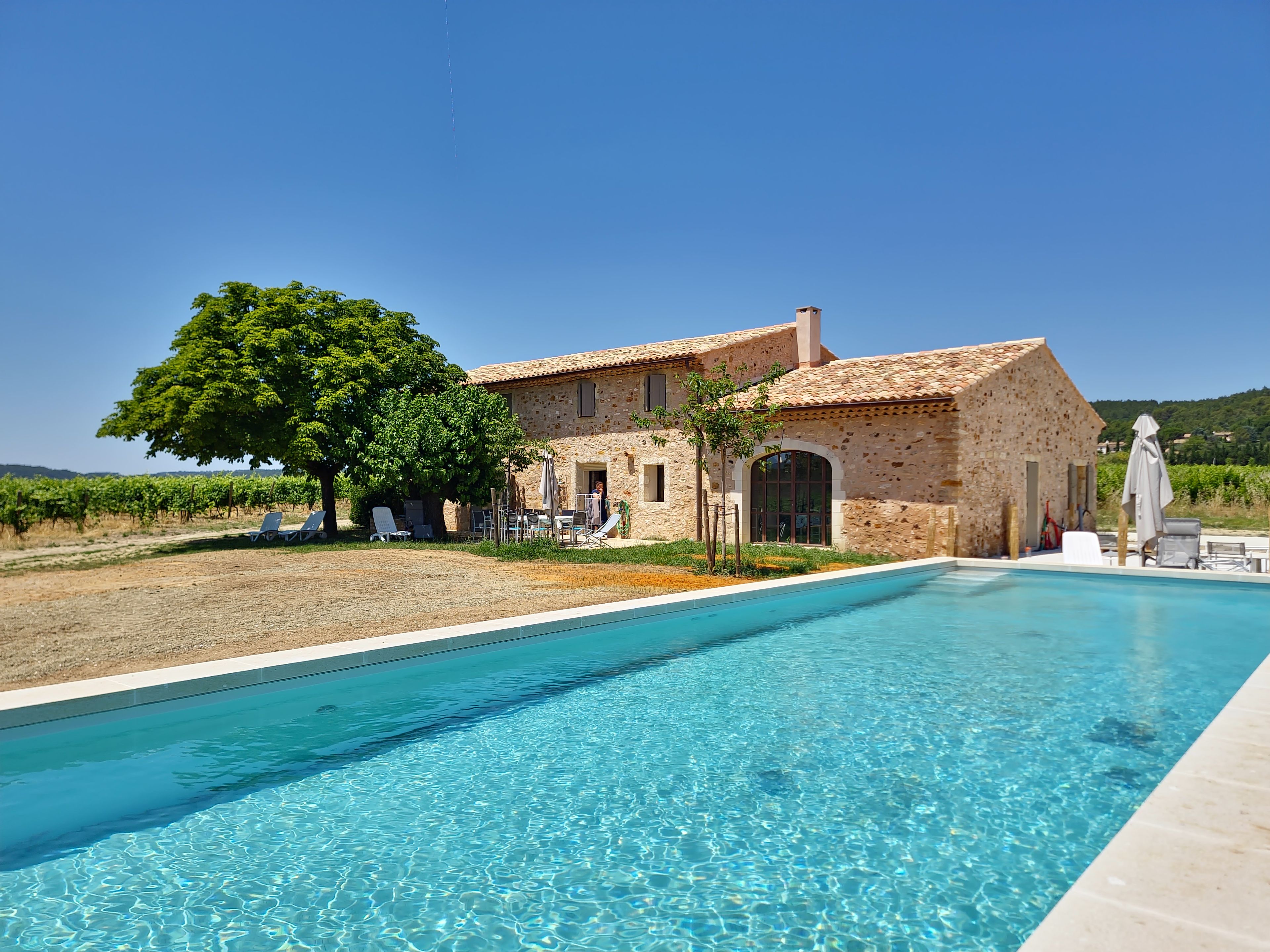 A poolside view of a stone building with a clear blue pool, outdoor seating, and a vineyard in the background.