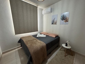 Ein Schlafzimmer mit Bett, Fenster mit Jalousie, Klimaanlage und einem kleinen Tisch mit einer Vase.