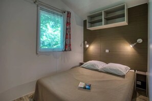 A bedroom with a bed, a window with curtains, a bedside table, and a shelf above the bed. - Camping 3 étoiles - Piscine  - ccbfgei (Groléjac)