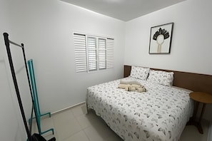 A bedroom with a bed, a window with blinds, a wall-mounted picture, and a door with a towel rack.