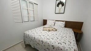 Quarto com uma cama, janela com persianas, quadro na parede e mesa de cabeceira.