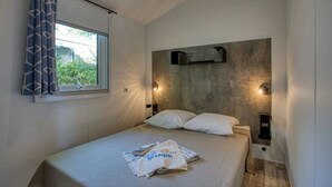 En enkeltseng med gavl, to vægmonterede lamper, et vindue med gardiner og et lille natbord med en bog og en taske.