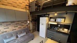 Um espaço de estar compacto com um sofá cinzento, cozinha com frigorífico e mesa de jantar.