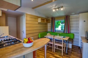 A compact kitchen with a gas stove, a bowl of fruit, and a dining area with a table and chairs. - Camping 3 étoiles - Piscine  - ccbfgfc (Groléjac)