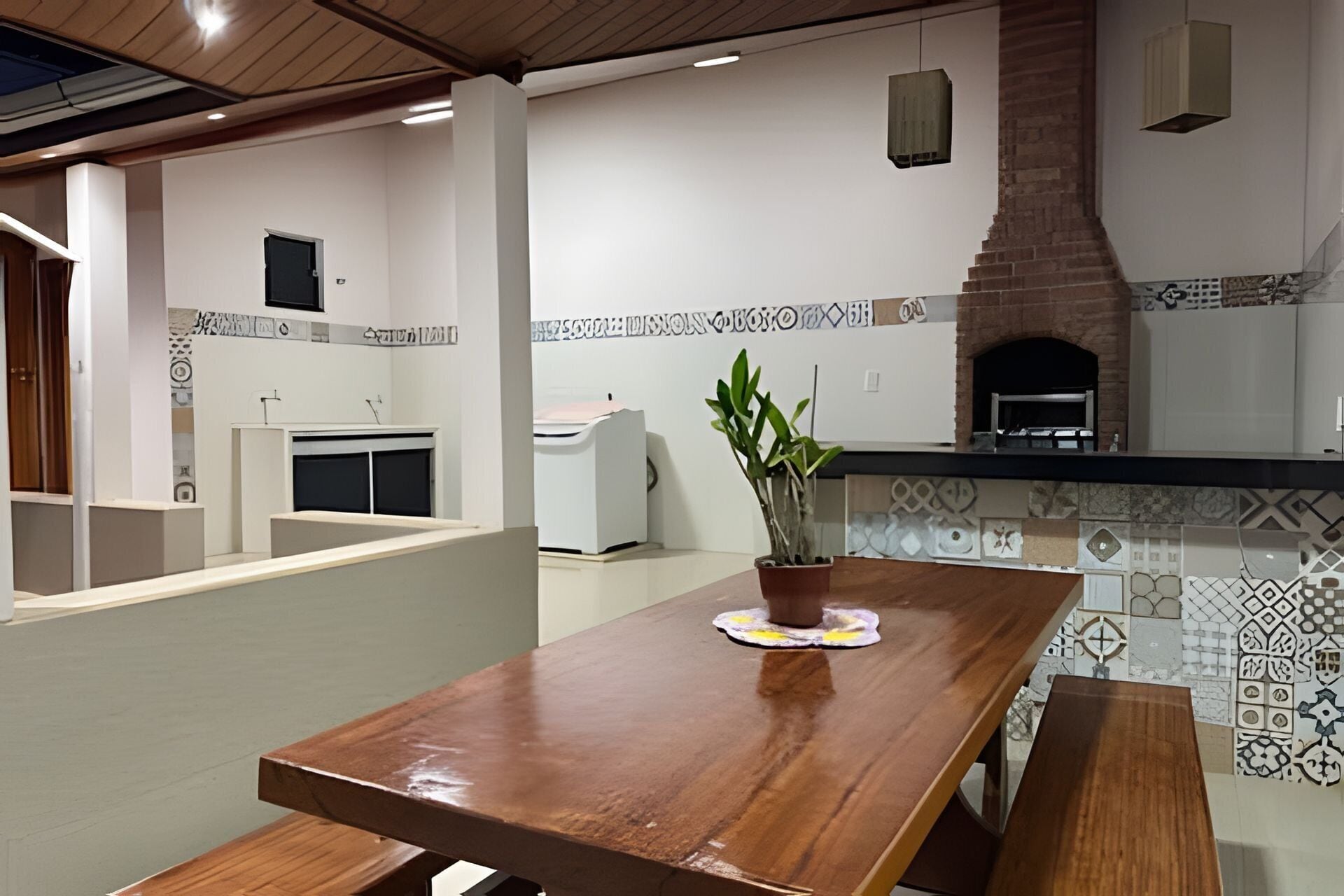 A wooden table with a potted plant, a kitchen area with a brick oven, and a patterned backsplash.