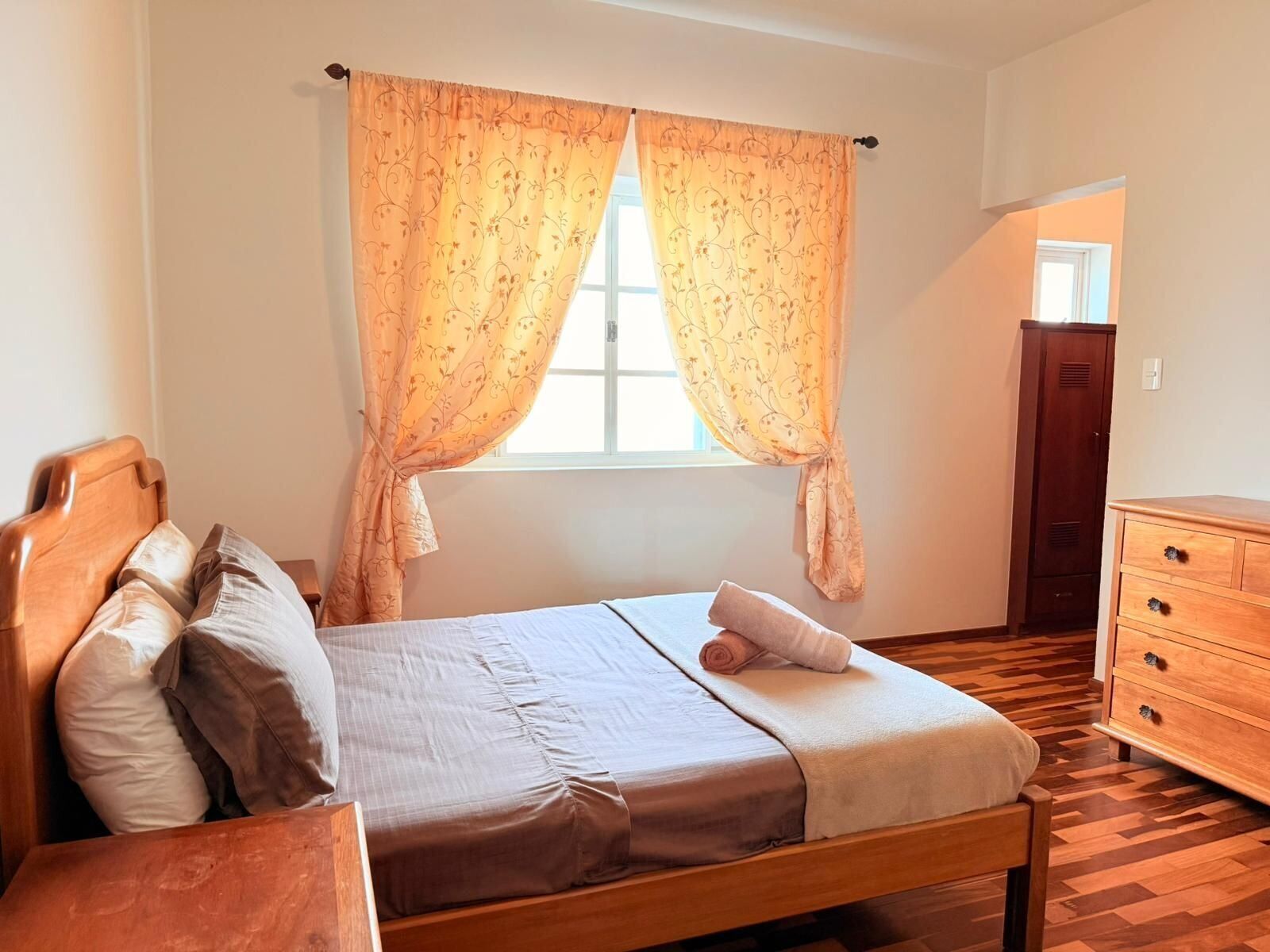 Ein Schlafzimmer mit einem hölzernen Bett, einer Holzkommode und einem Fenster mit Vorhängen.