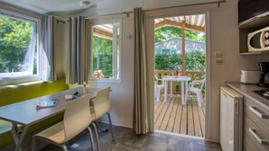 A room with a dining area, a kitchen, and a view of a wooden deck and greenery. - Camping 3 étoiles - Piscine  - ccbfgff (Groléjac)