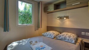 A bedroom with a bed, pillows, a window, curtains, and a wall-mounted shelf. - Camping 3 étoiles - Piscine  - ccbfgff (Groléjac)
