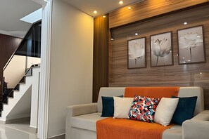 A modern living room with a staircase, a sofa with colorful cushions, and three framed wall art pieces.