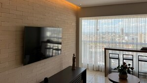 Ein modernes Wohnzimmer mit Flachbildfernseher an einer Ziegelwand, einem Essbereich mit rundem Tisch und einem großen Fenster mit Blick auf die Stadt.