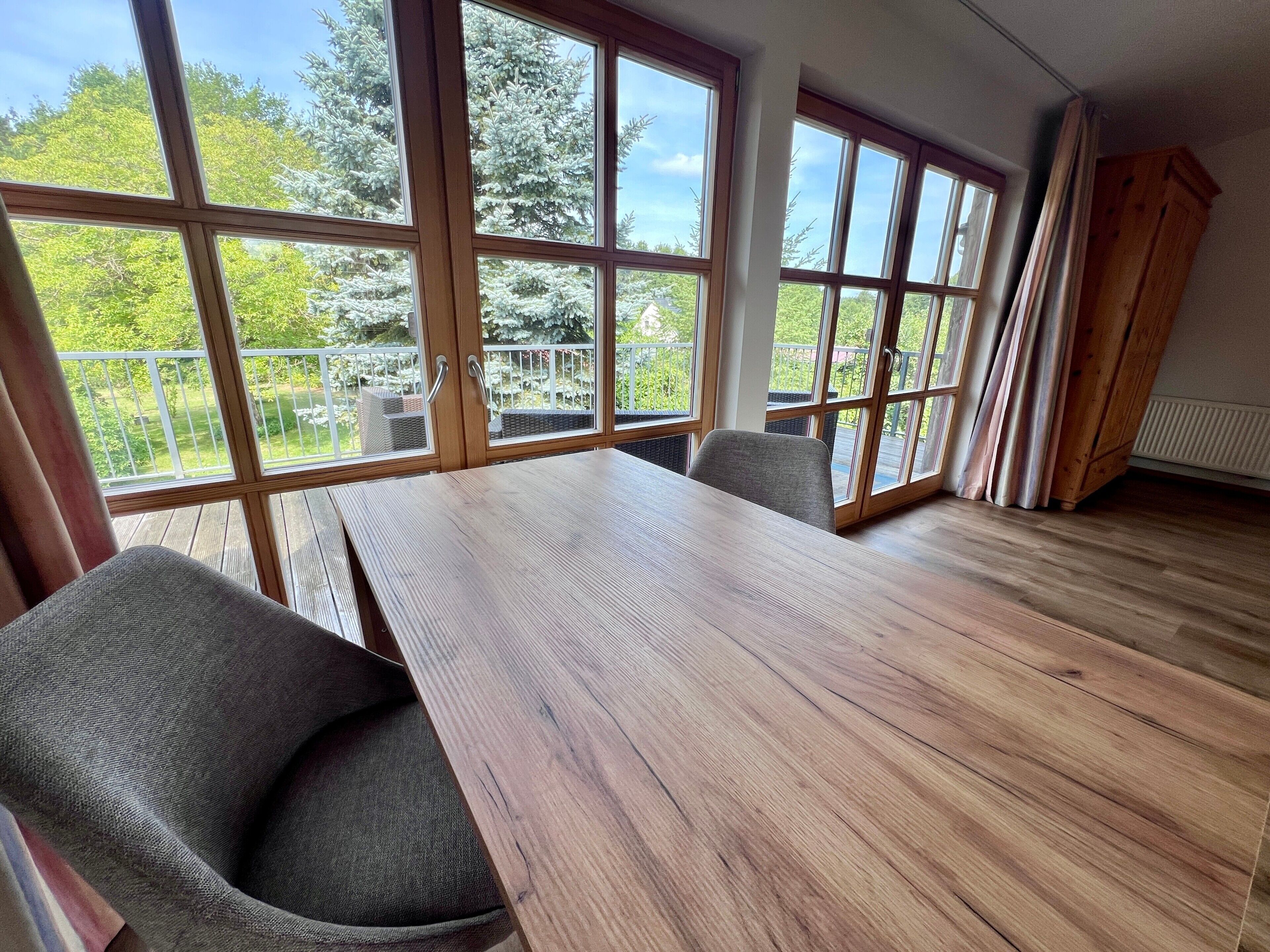 A room with a large wooden table, two chairs, and a view of a balcony with a railing.