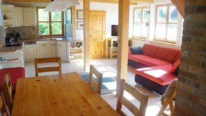 A wooden dining table with chairs, a red sofa, a kitchen with white cabinets, and a window with a view of greenery.