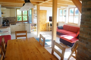 Een houten eettafel met stoelen, een rode bank, een keuken met witte kastjes en een raam met uitzicht op groen.