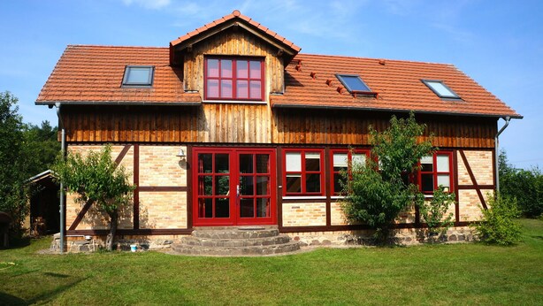 A two-story wooden house with a red door, multiple windows, and a sloped roof.