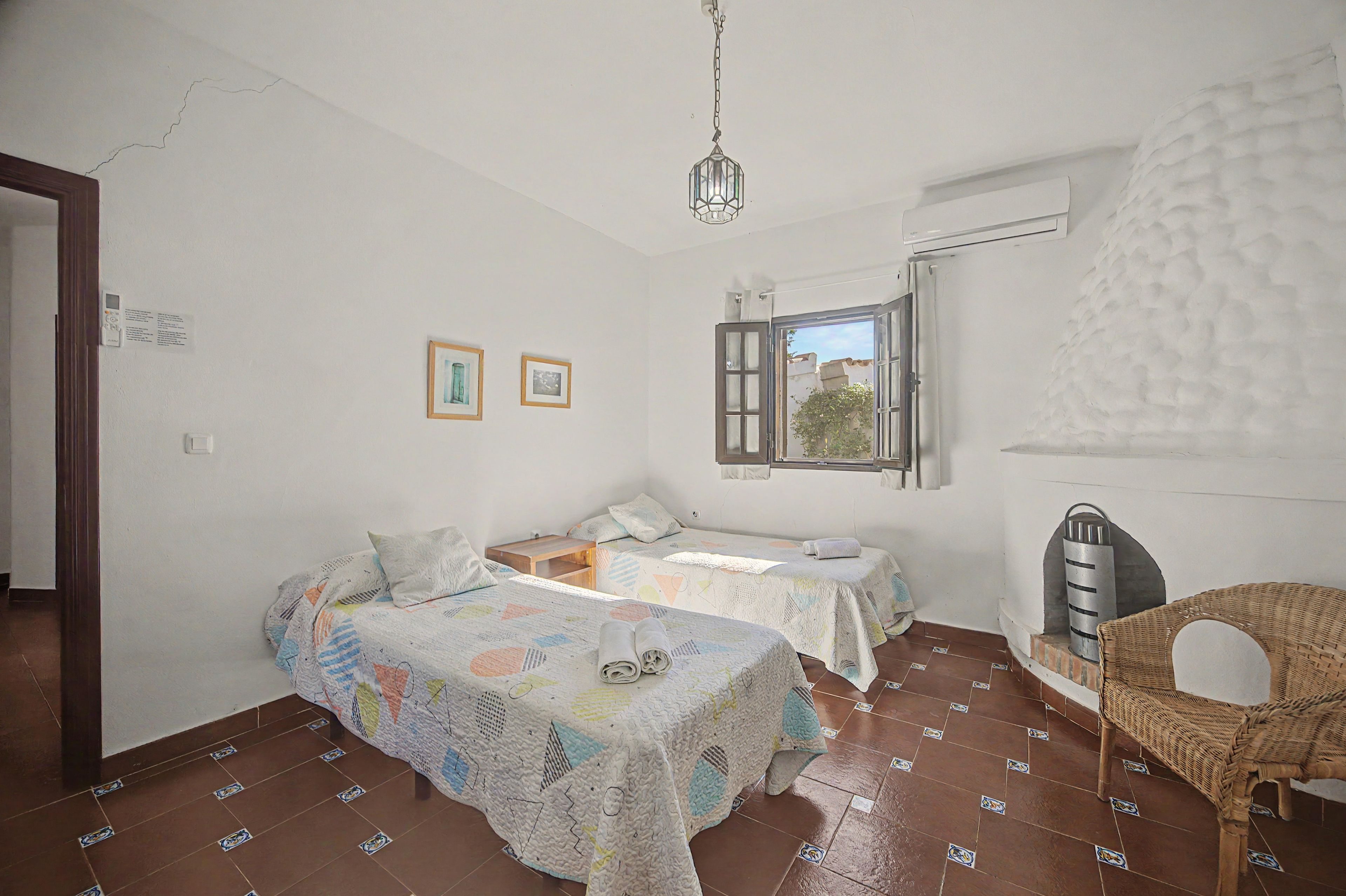 A bedroom with two beds, a window, a hanging light fixture, and a wicker chair.