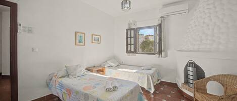 A bedroom with two beds, a window, a hanging light fixture, and a wicker chair.