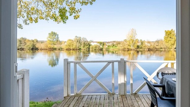 Een houten terras met een witte balustrade, uitkijkend op een rustig meer met bomen op de achtergrond.