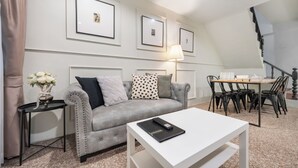 Una sala de estar con un sofá gris, una mesa de centro blanca, una mesa de comedor con sillas, cuadros enmarcados en la pared y una escalera.