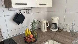 Une cuisine avec des meubles blancs, une machine à café, un panier de fruits et une bouilloire.