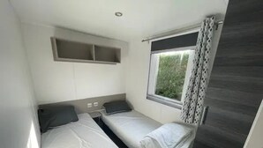 A compact room with two beds, a window with curtains, a shelf above the beds, and a door. - Camping 4 étoiles - Parc aquatique - ccbfgac (Les Mathes)