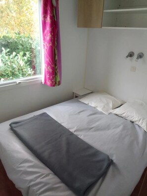 A single bed with a gray blanket, white pillows, a small bedside table, a window with a pink curtain, and a wall-mounted shelf.