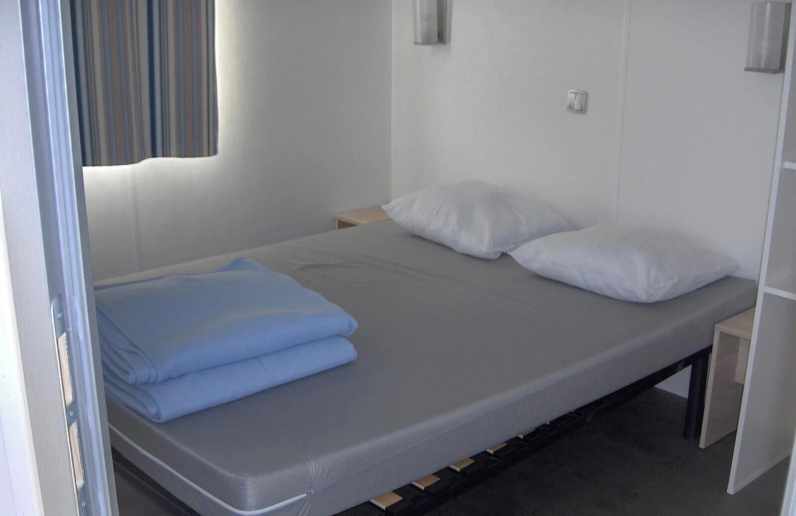 A single bed with a grey mattress, white pillows, and a blue blanket. There is a small bedside table with a lamp, and a wardrobe is visible to the right.