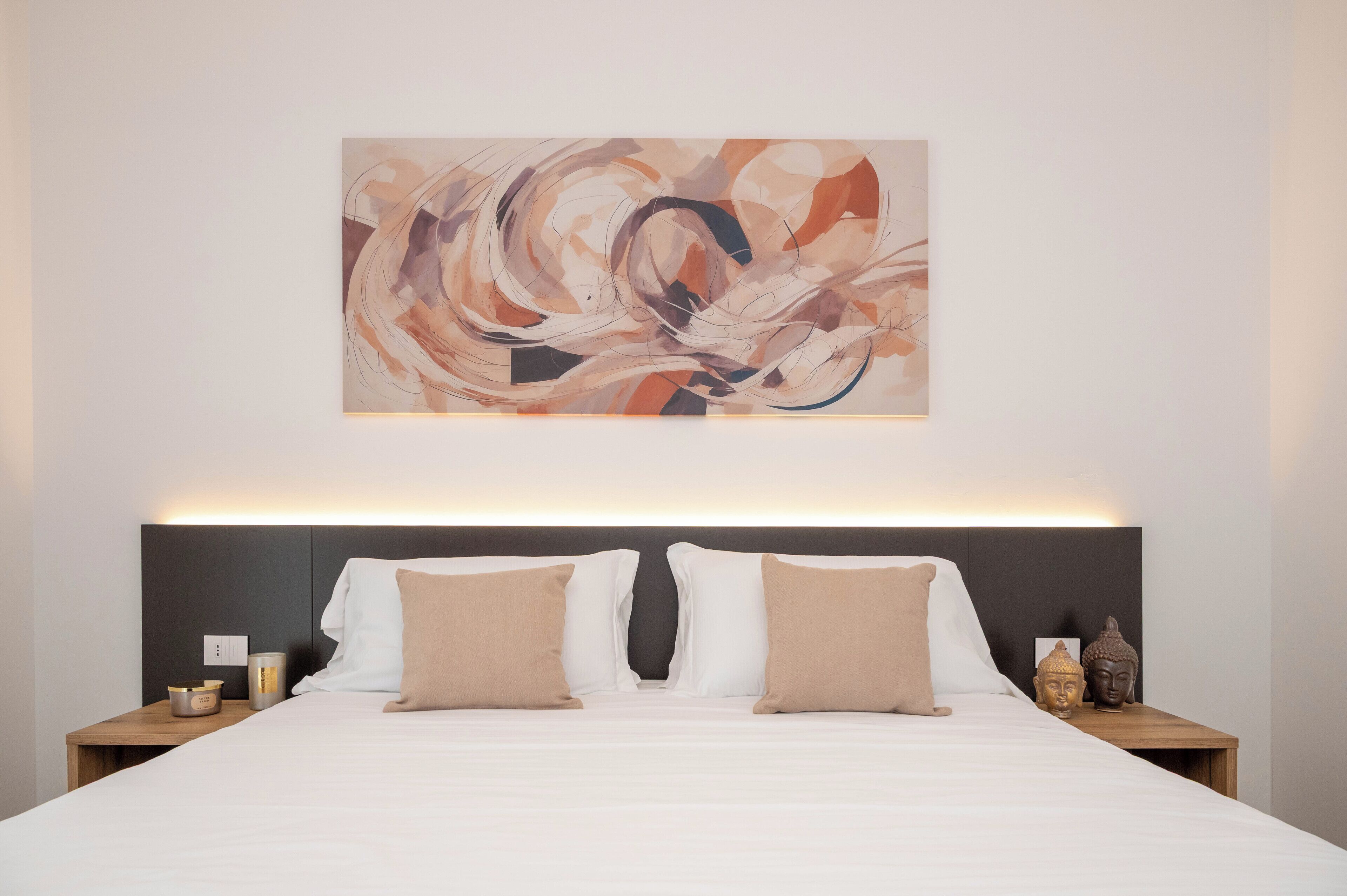 A modern hotel room with a large abstract painting, a neatly made bed with two pillows, a bedside table with a lamp, and a small Buddha statue.