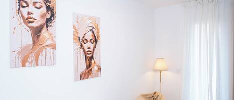 Quarto com duas pinturas na parede, uma luminária de cabeceira, uma janela com cortinas e uma cama.