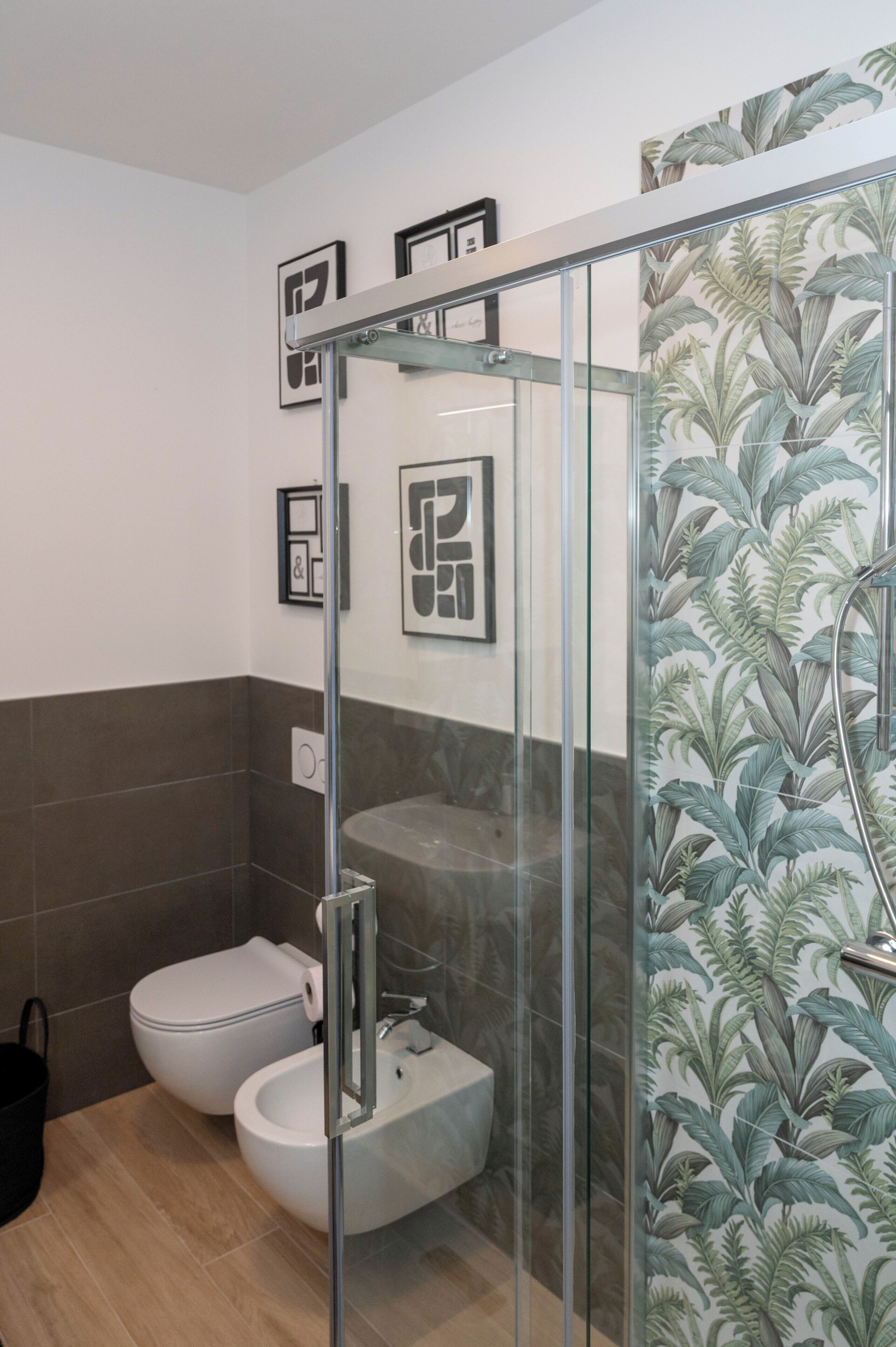 A bathroom with a glass shower enclosure, a toilet, and a bidet. There are framed pictures on the wall and a tropical leaf-patterned wallpaper.