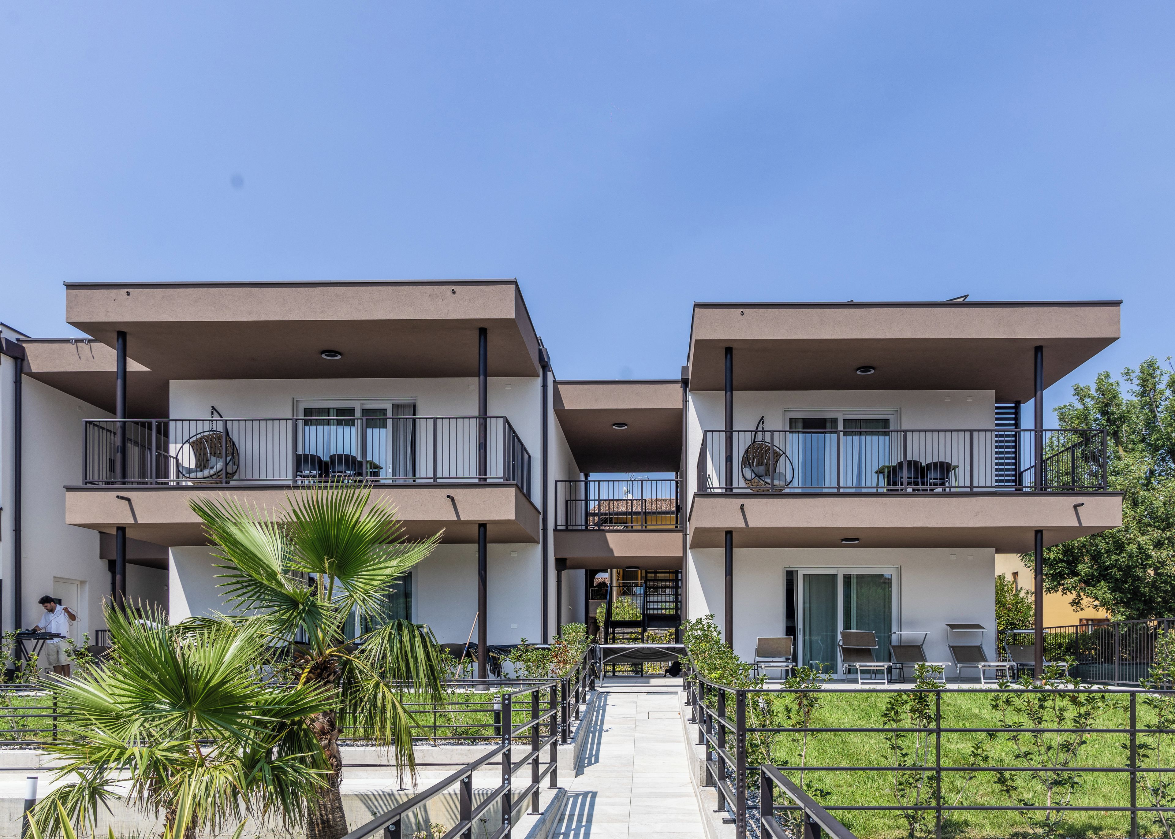 A modern two-story residential building with balconies, outdoor seating, and a pathway leading to the entrance.