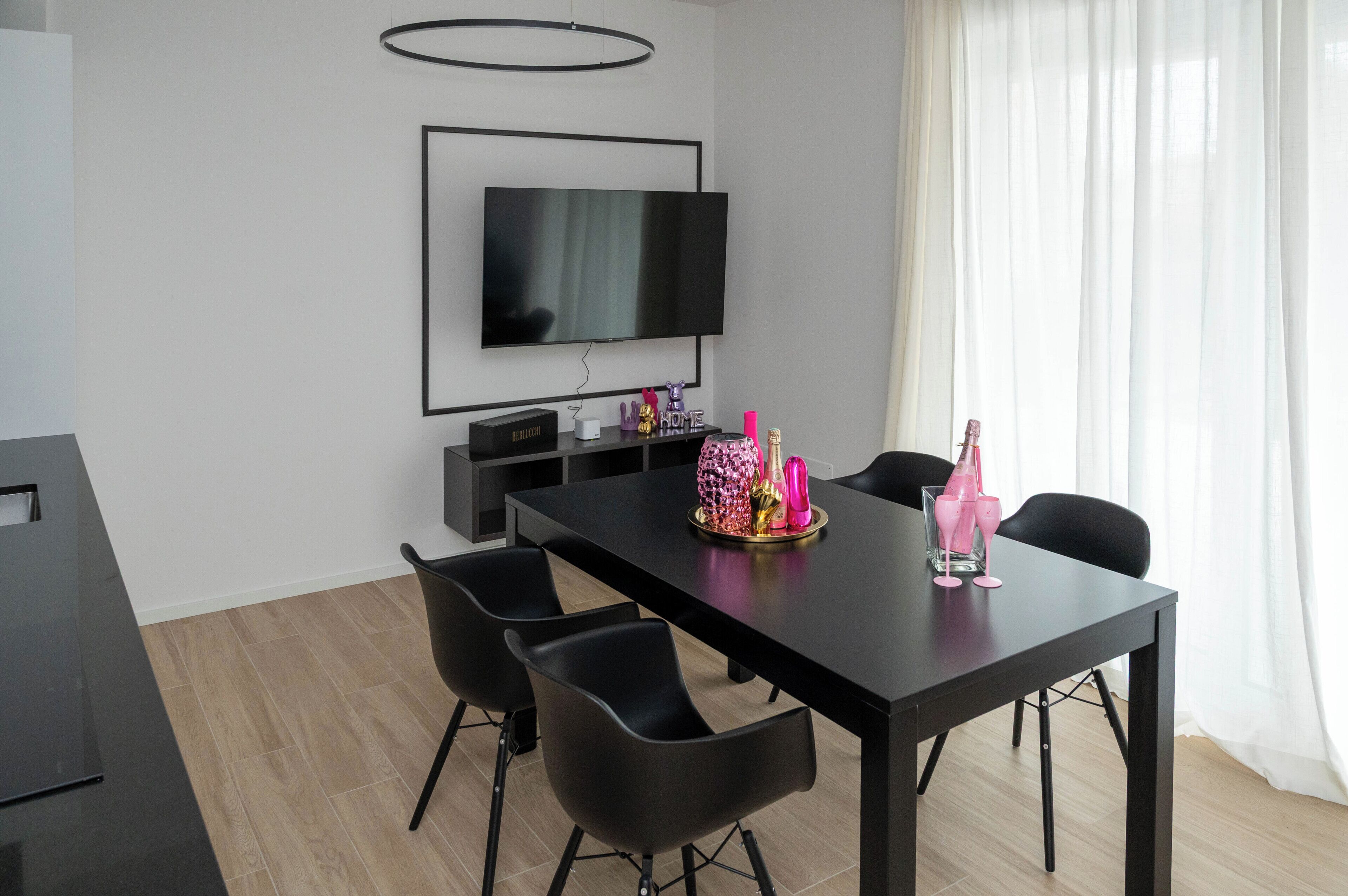 Un espace repas moderne avec une table noire, des chaises noires, un téléviseur à écran plat fixé au mur et un plateau décoratif contenant des objets roses et dorés.