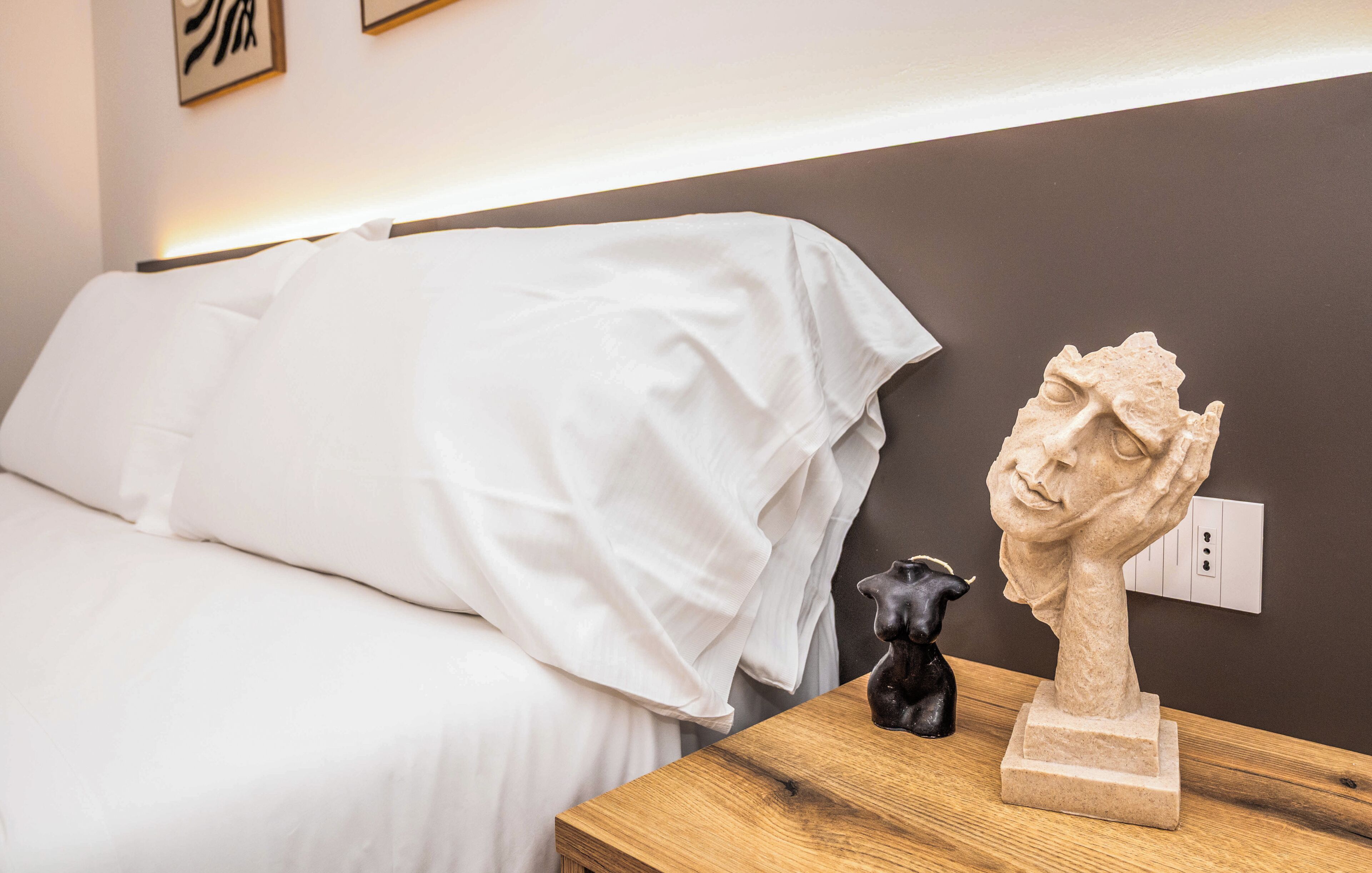 A bed with white bedding, a wooden nightstand with a sculpture, and a wall socket.