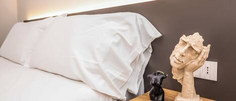 A bed with white bedding, a wooden nightstand with a sculpture, and a wall socket.