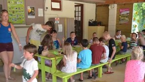 Ein Klassenzimmer mit Kindern, die an grünen Pulten sitzen, einer stehenden Lehrerin und einem Kind, das geht.