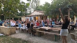 Draußen versammeln sich Menschen an Tischen, im Hintergrund ist ein Gebäude zu sehen und eine Person steht mit erhobenen Armen.