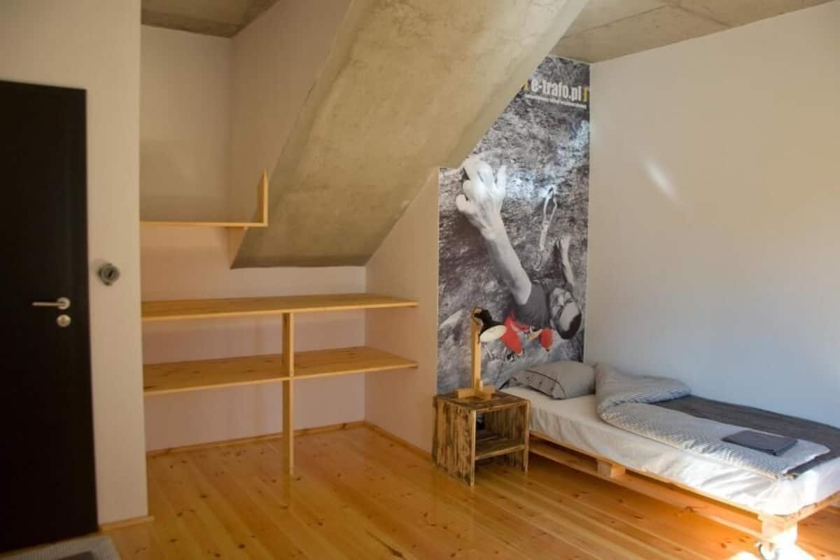 A room with a sloped ceiling, a bed with a headboard, a small wooden nightstand, and a wall-mounted shelf.