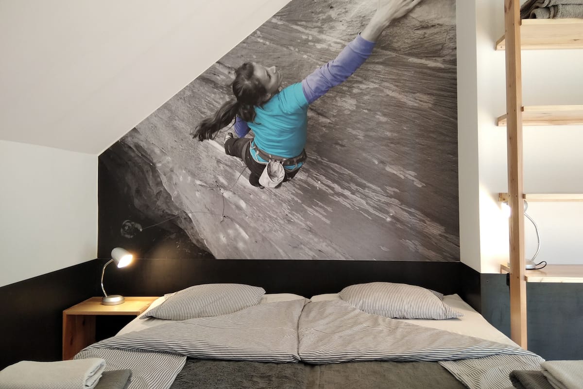 A bedroom with a bed, a nightstand, a lamp, a wall with a climbing wall mural, and a wooden shelf.