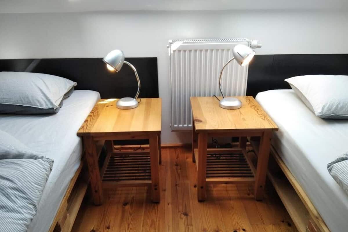 A bedroom with two beds, each with a wooden nightstand and a desk lamp. A radiator is visible in the background.