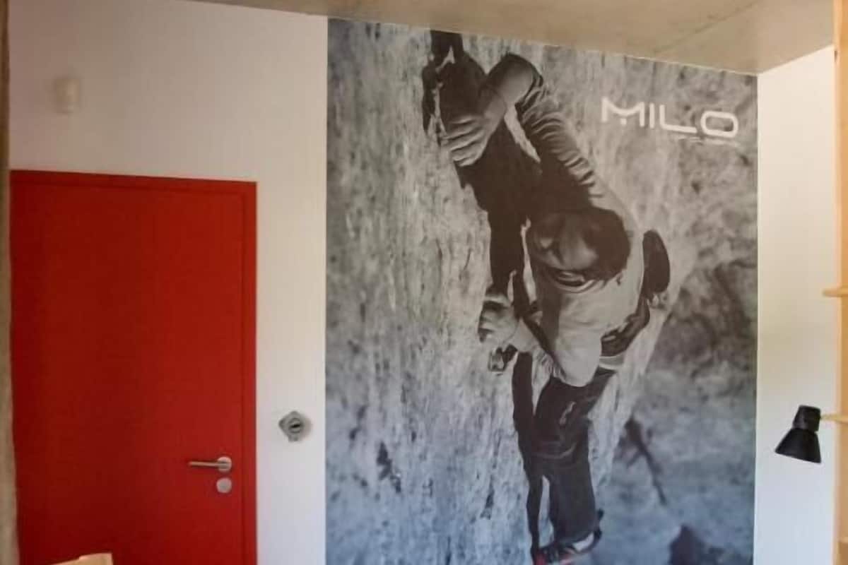 A room with a red door, a bed, a wooden table with chairs, and a wall mural of a person climbing.