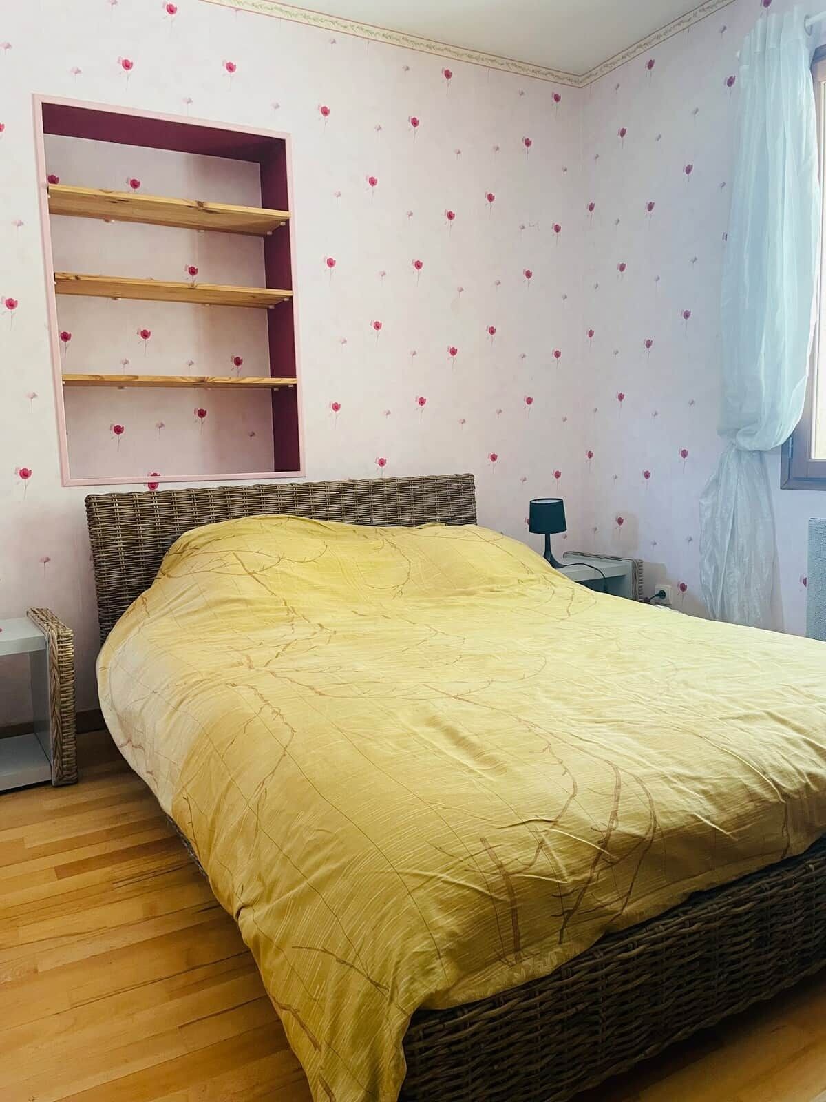 A bedroom with a bed, a shelf unit, a bedside table, and a window with curtains.