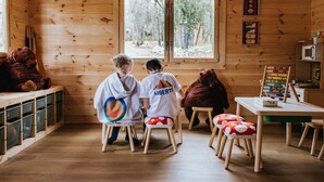 Interior de uma cabana de madeira com duas crianças em banquinhos, um ursinho de pelúcia e uma mesa com micro-ondas.