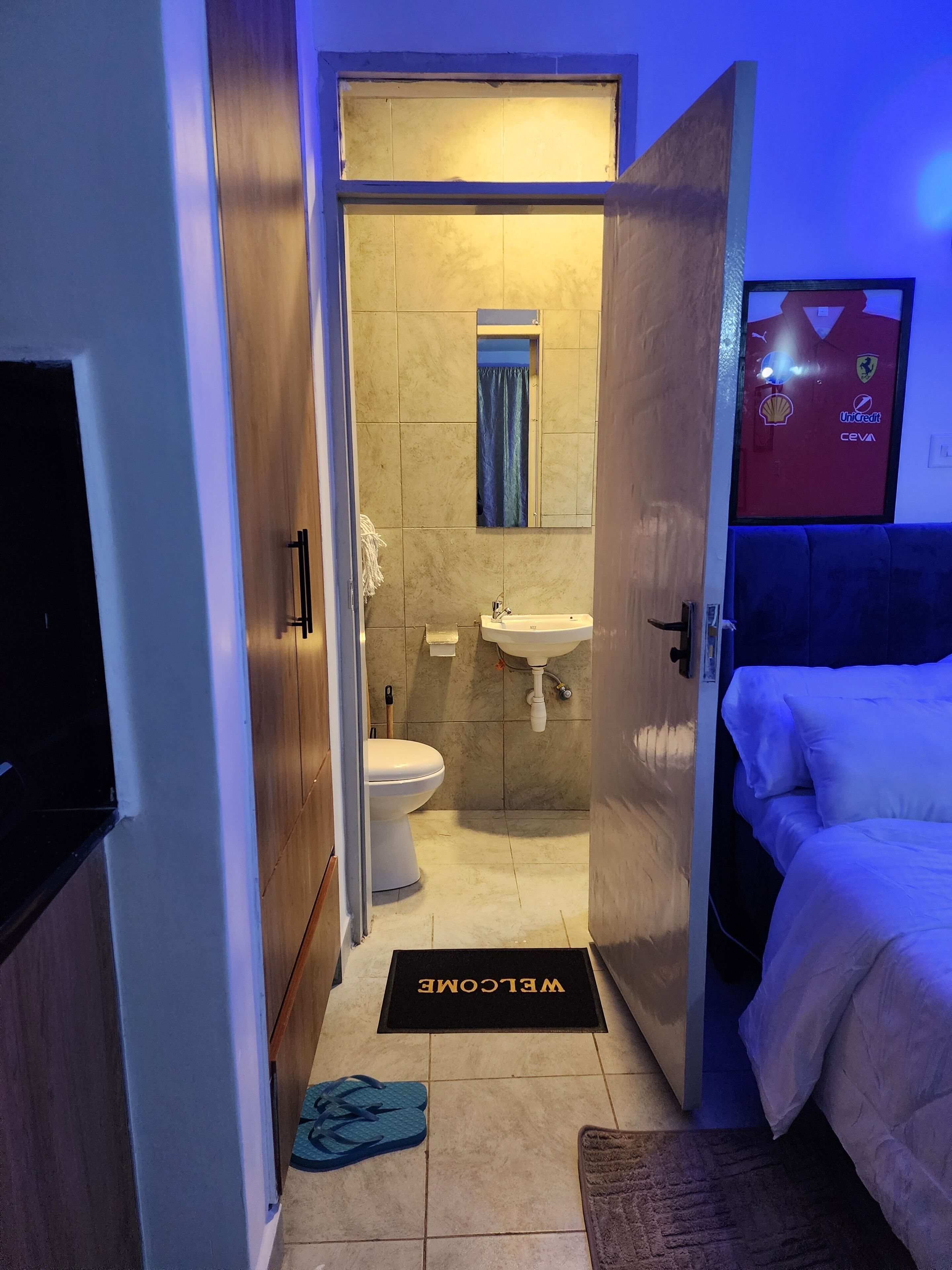 A hotel room with a bed, a bathroom with a toilet and sink, a welcome mat, and a wall-mounted TV.