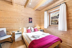 Un dormitorio de cabaña de madera con una cama, mesitas de noche, una silla y una ventana con cortinas.