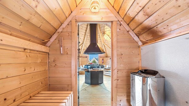 Innenansicht einer Holzhütte mit Kamin, einem Metallheizkörper und Blick durch eine Tür in einen Außenbereich.