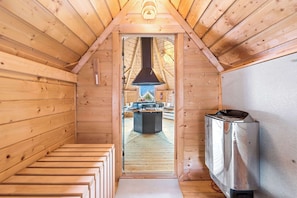 Interior de una cabaña de madera con chimenea central, un calentador metálico y vistas al exterior desde la puerta.