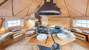 Ein Holzblockhaus im Inneren mit einem zentralen Feuerkorb, Holzbänken und einer hohen Decke.